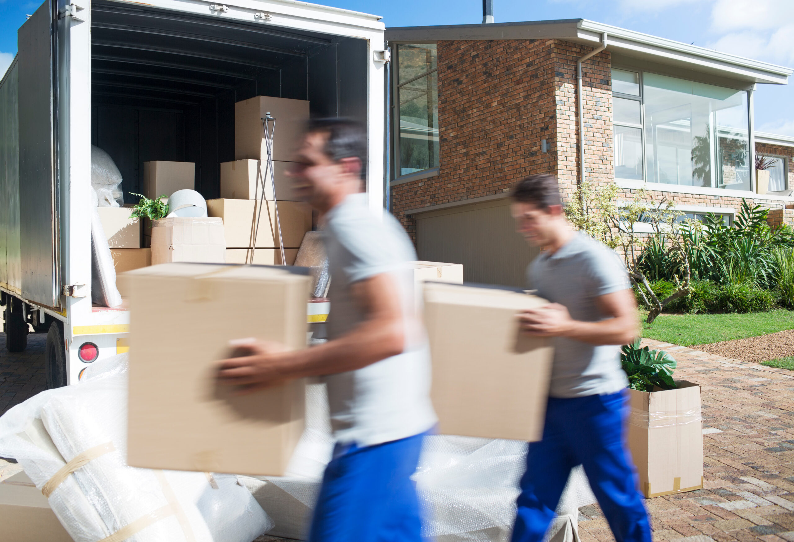 Two professional movers in uniform carrying cardboard boxes from a moving truck at a residential home